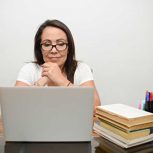 A middle-aged woman with dark brown hair and glasses is seated and looking at a laptop. A glass of lemonade is sitting on the desk to her left. To her right is a stack of books and an assortment of colorful pens.
