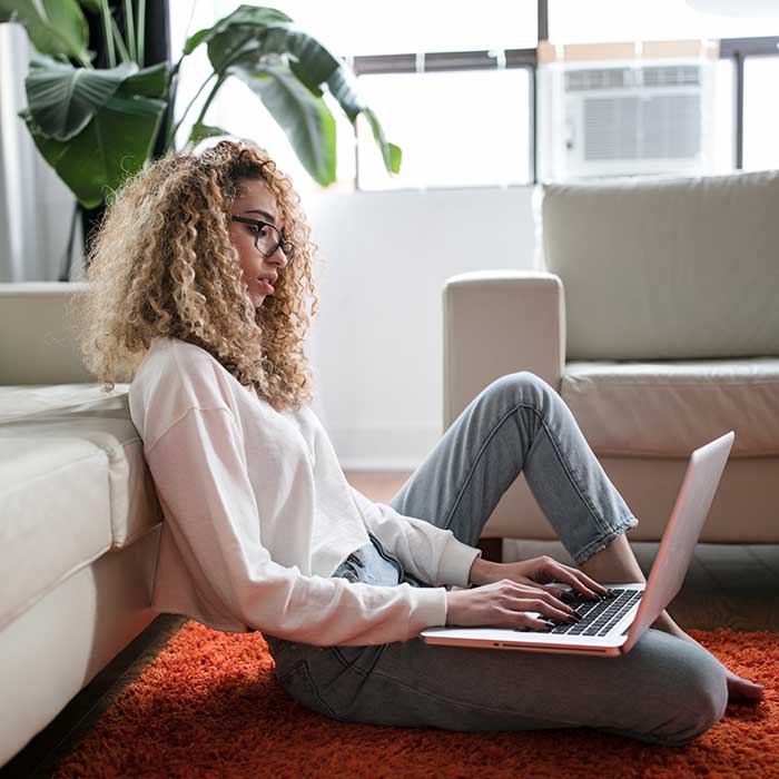 Woman sitting on the floor, working on a laptop, leaning against a couch. 