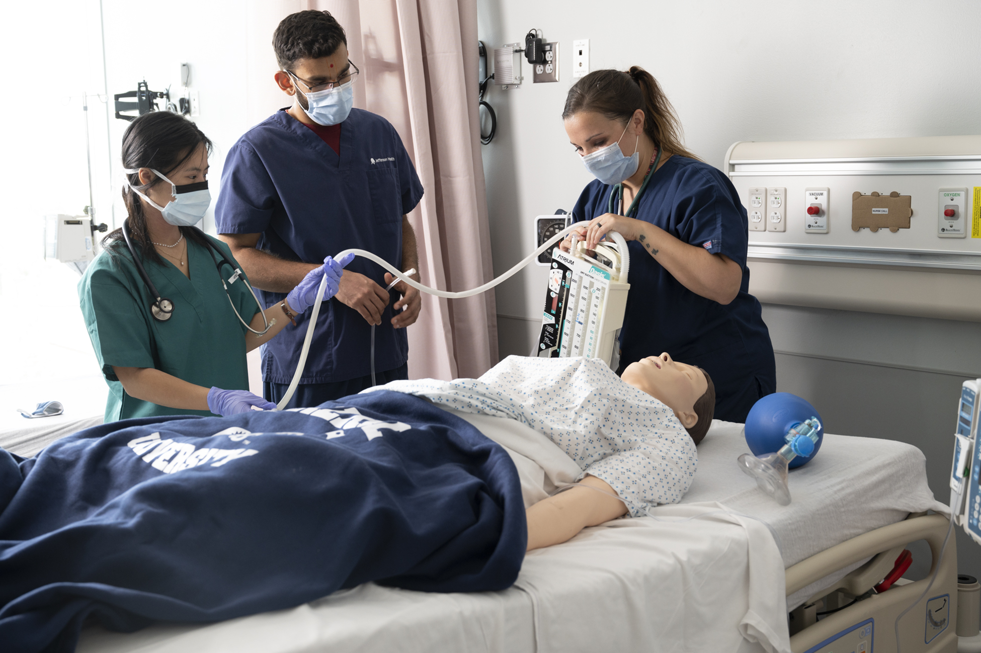 three health professionals surround manikin laying in hospital bed
