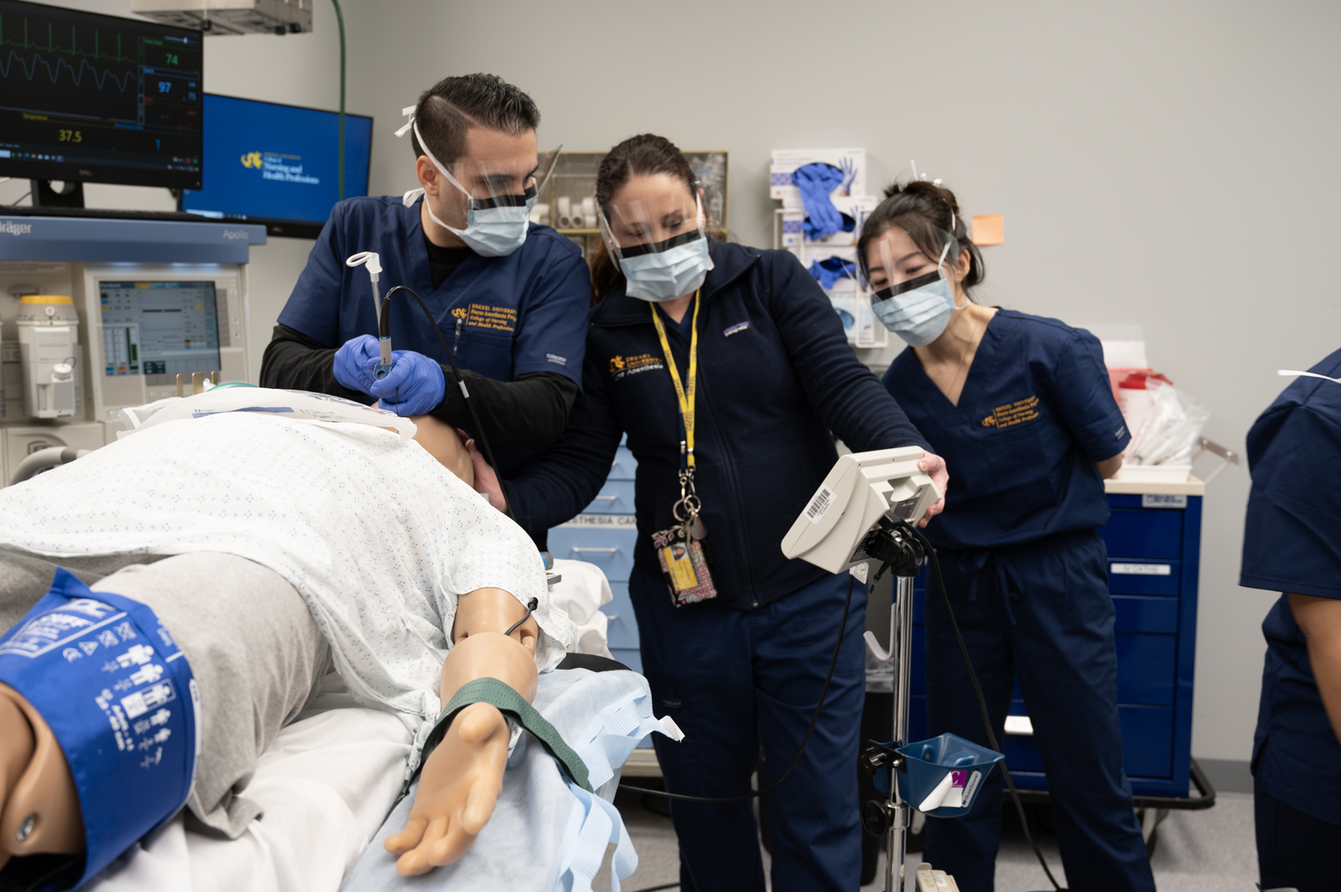 Three health care professionals look at operating room equipment, manikin on operating table