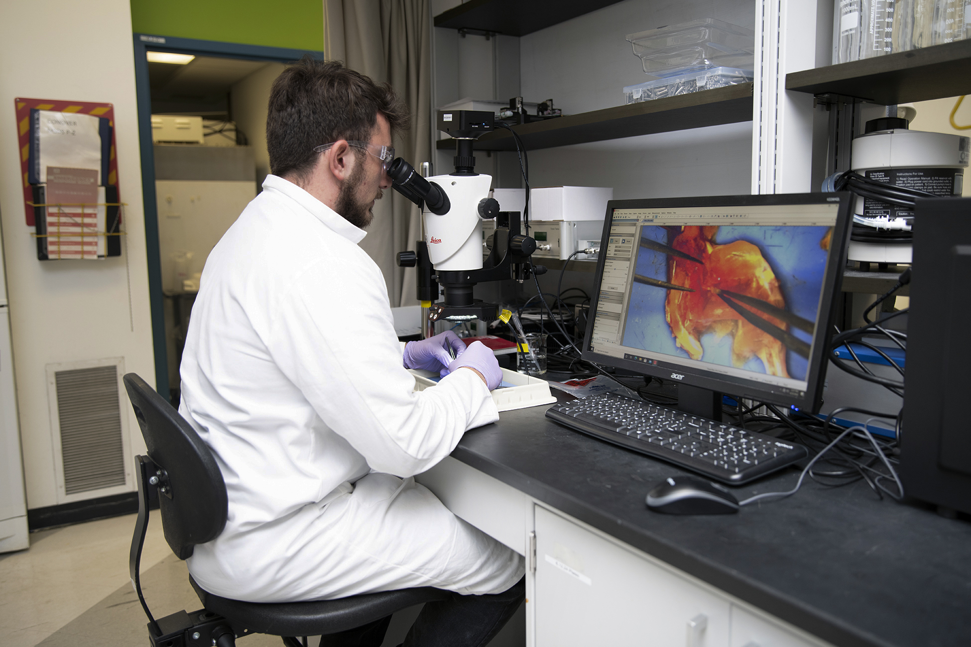 Man in white lab coat looks through microscope with image projected on computer screen