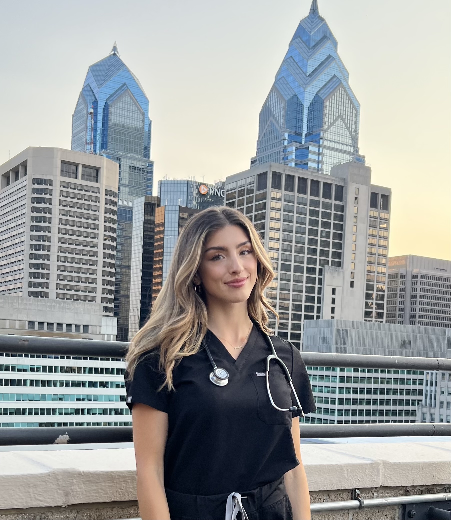 PA Student Cassie Job wearing black scrubs with stethoscope draped around her neck, long blond hair over her shoulders, standing on a bridge with Philadelphia city skyline in the background