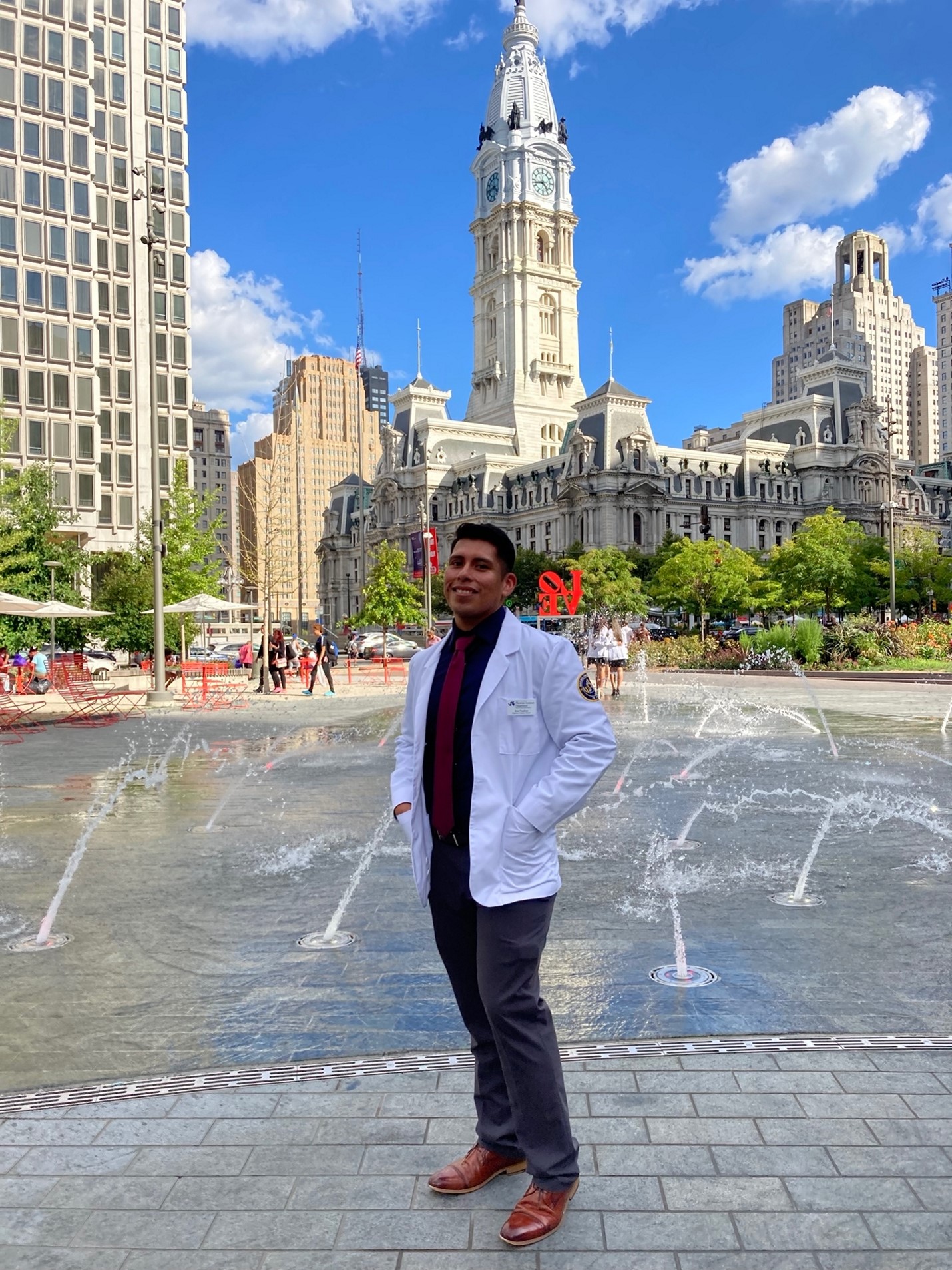 PA Student Jose Cuadros wearing white coat, standing by fountains in Love Park with Philadelphia City Hall in the background