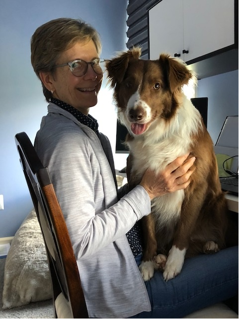 Julie Kinzel sits in a desk chair smiling with a large collie dog in her lap