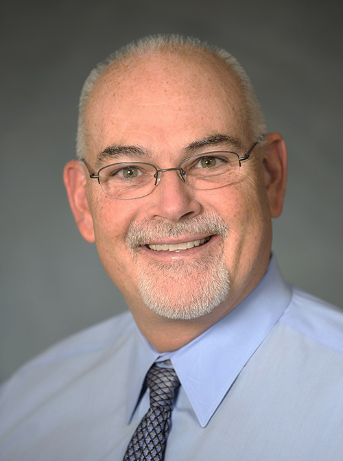 headshot of preceptor Andrew Kelly wearing glasses, white goatee, blue collared shirt and blue tie