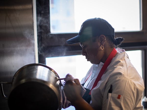 Chef Tonii Hicks, a Black female, is wearing a white chef's coat and baseball cap, pouring steaming contents of a large pot.