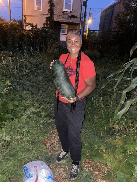 Chef Tonii Hicks, a Black female wearing a red tshirt and dark pants, is holding a large melon in a garden at night.