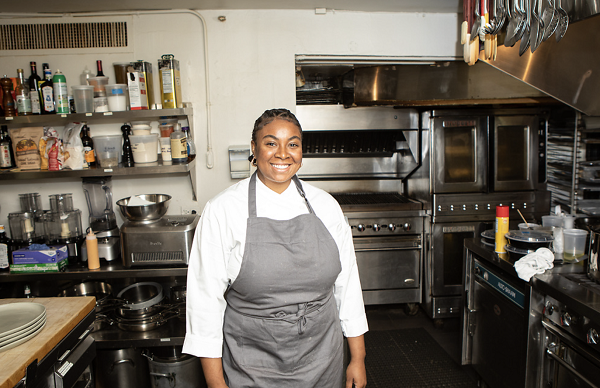 Chef Tonii Hicks, a Black female, is wearing a white chef's coat and gray apron and standing in the middle of a professional kitchen.
