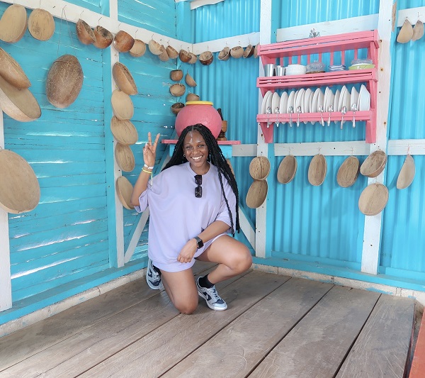 Young Black woman with long braids and lavender shirt kneeling in the corner of a room with light blue walls adorned with baskets