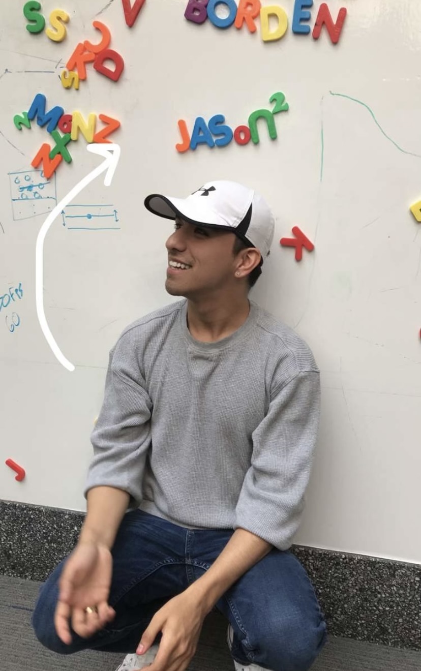 Master's of Art Therapy and Counseling student Jason Valdez squatting in front of a white board with alphabet letters spelling his name behind him..
