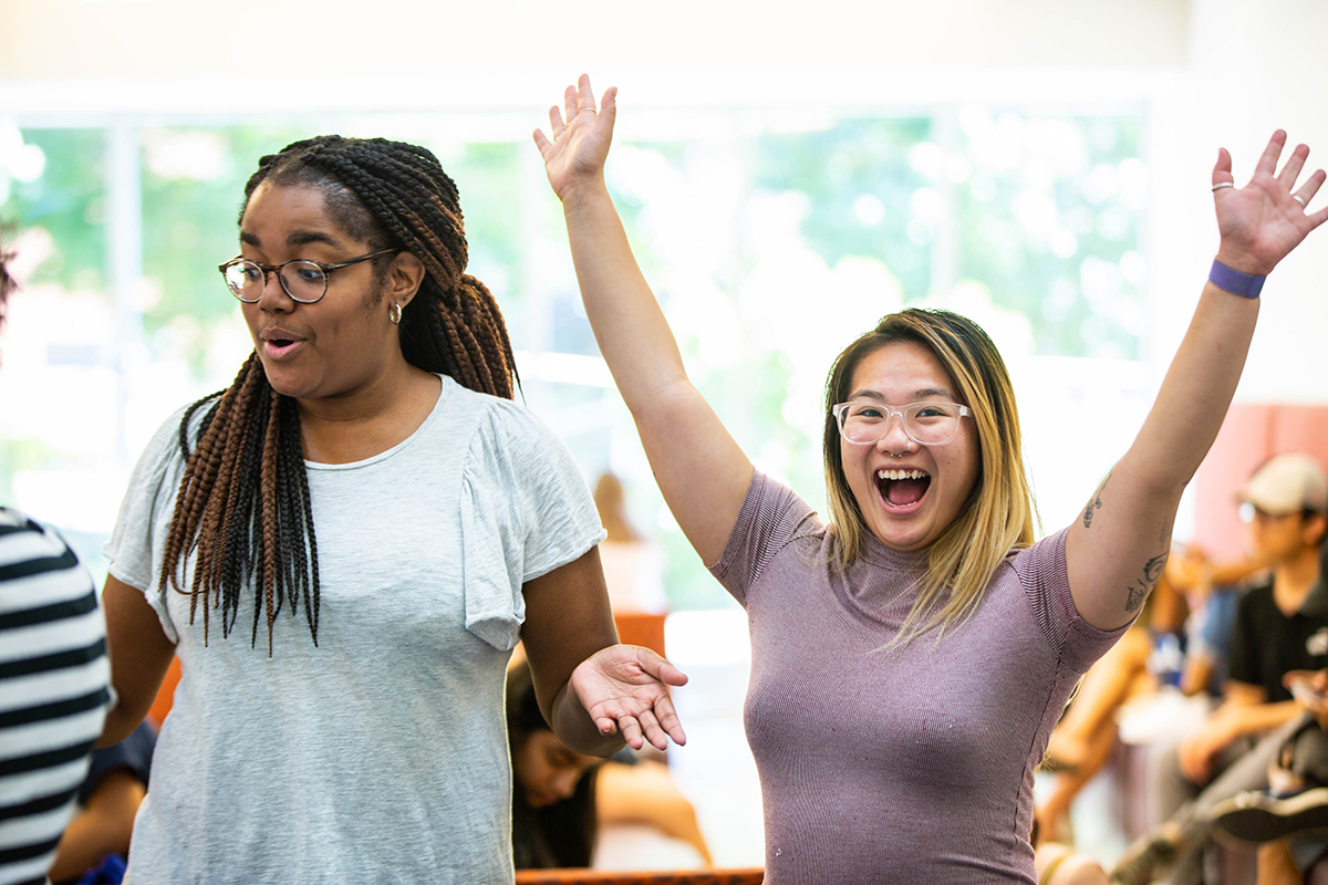 Two students are at an event. Both are smiling and one student has her arms raised above her head in celebration.