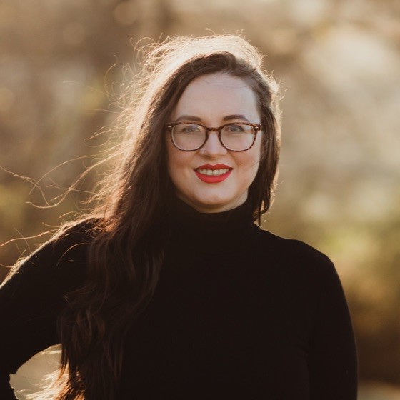Mary Madsen smiles outside during golden hour, wearing a black turtleneck and glasses