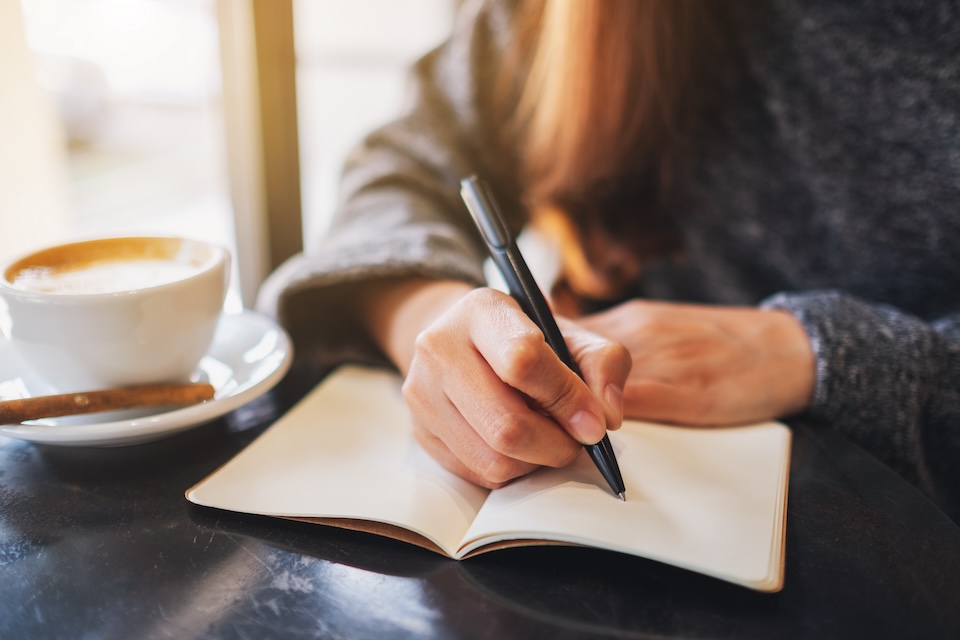 A person writes in a notebook beside a cup of coffee
