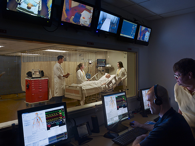 Three nursing students interact at the bedside of a simulated patient while two instructors observe remotely from a control room