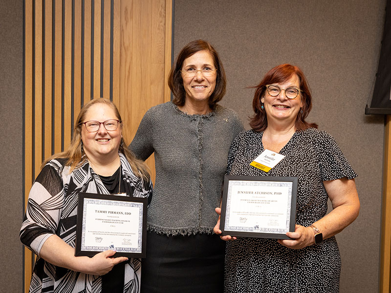 Two women holding award certificates presented by a third woman