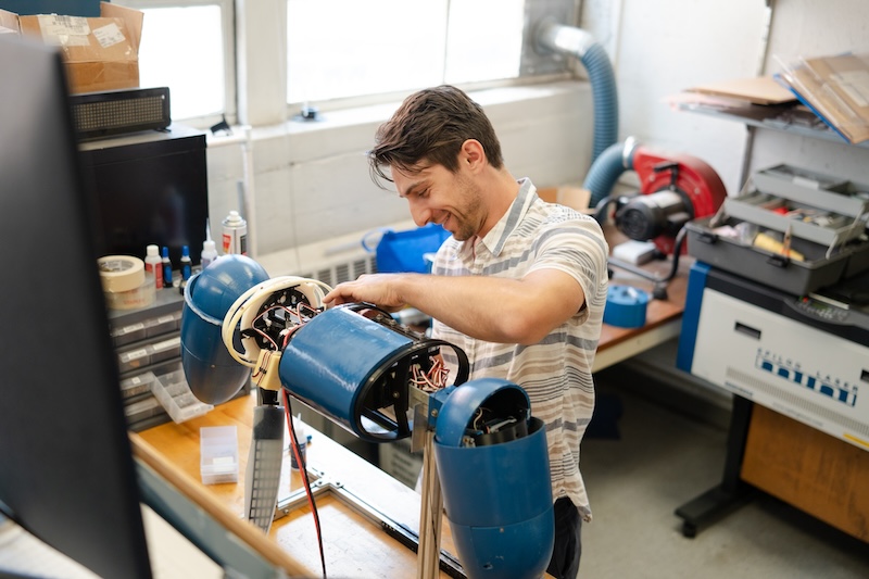 A student stands at a work table, adjusting the electronics on a blue, cylindrical robot shaped like a sea lion.