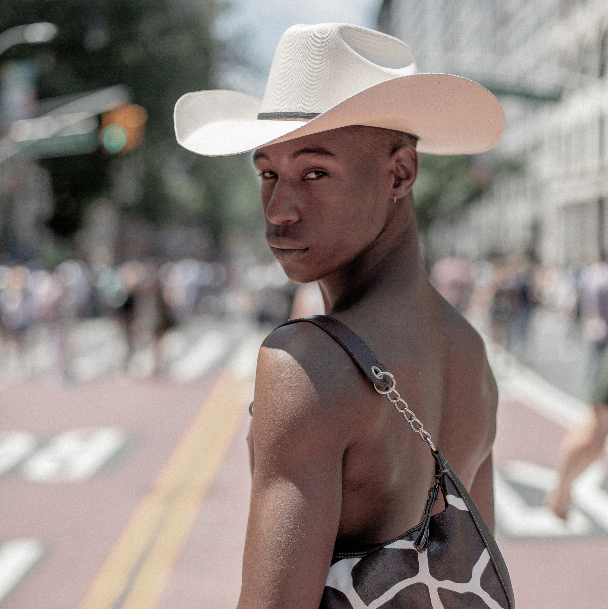 Fashionable young person of color looking over their shoulder. They are shirtless with a cowboy hat and a purse slung over their shoulder.