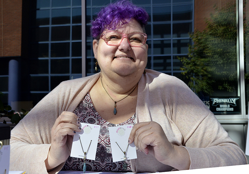 Woman with purple hair and glasses smiling while holding up handmade necklaces