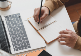 Hands writing in notebook with laptop and coffee nearby