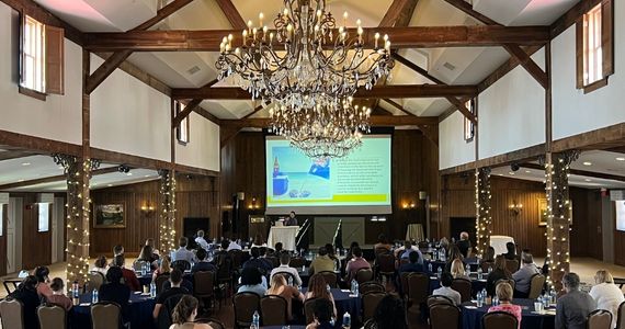 Conference room with large chandelier and people seated at roundtables facing a large screen with a presentation 