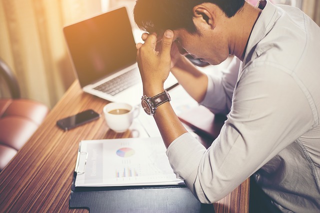 Image of a man frustrated as he leans over his paper, with an open laptop to his right.