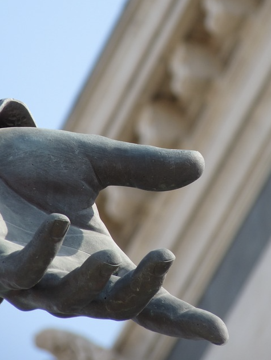 Image of a bronze statue hand with the cornice of an ornate building in the background