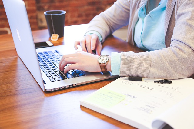 Image of a person sitting at a computer in work clothes with papers and a cup of tea next to them.