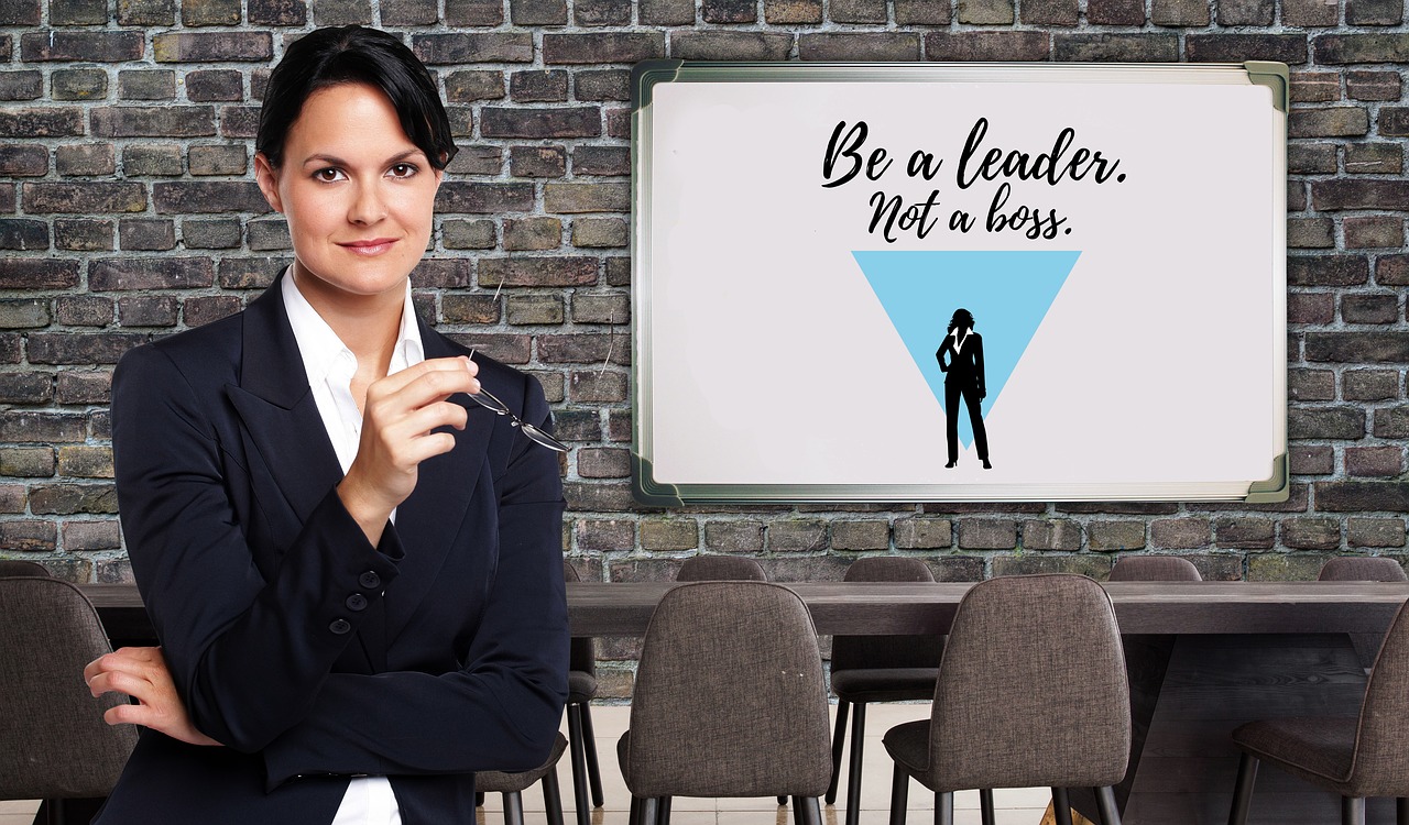 Image of a woman with dark hair in a suite holding a pair of eye glasses with the white board to her right that states: "Be a leader, not a boss."