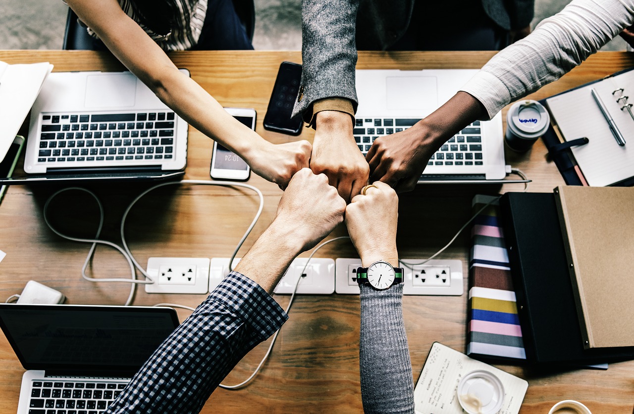 Image of a group of co-workers reaching across a table with computers in a fist pump, as in being supportive of one another.