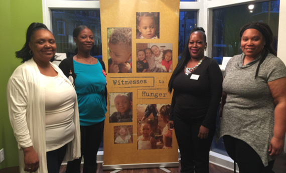 Four female members of Witnesses to Hunger pose next to their program banner