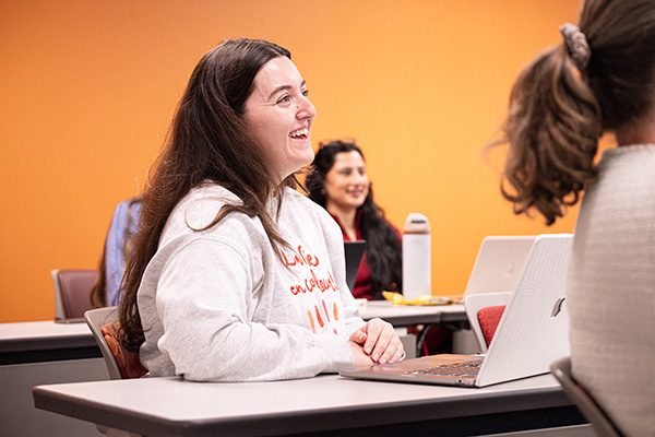 A student sitting at a desk in a classroom smiles.