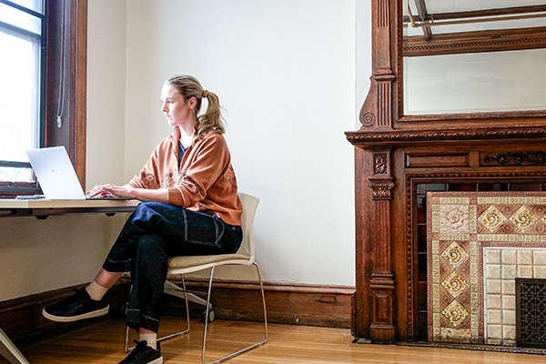 A student typing on a laptop while sitting near a fireplace in the Dornsife Center for Neighborhood Partnerships.