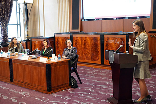 At the conference, a woman speaks at a lectern while a panel of three women look on. 