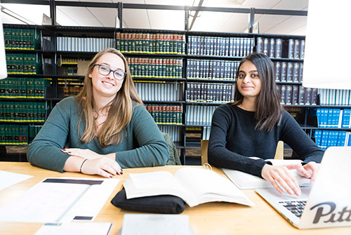 Two female law students facing camera while sitting at table in library with shelves full of books behind them.