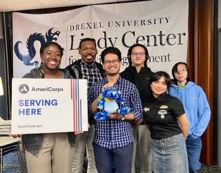 A group of 6 people stands together smiling with a sign that says 'AmeriCorps Serving Here'. The people have a variety of skin colors, hair colors, and clothing. Behind them is a banner for the Lindy Center for Civic Engagement.