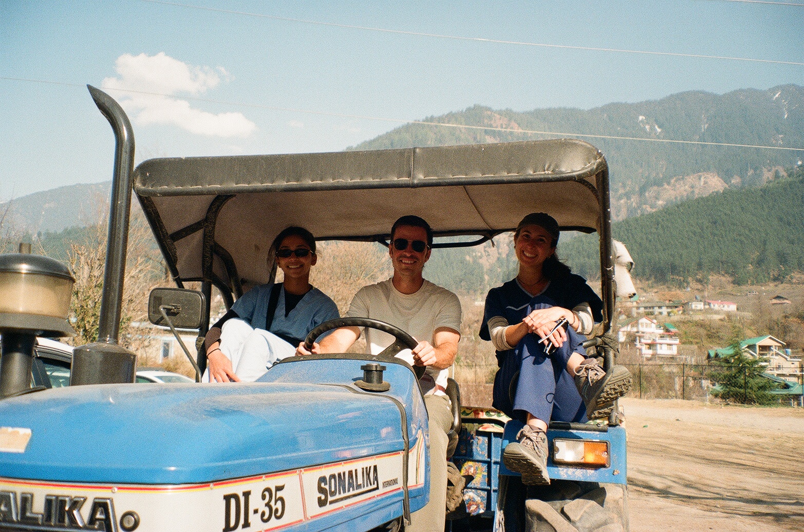Deeksha Ajeya riding a tractor with two others on a dirt road with mountain terrain in the background