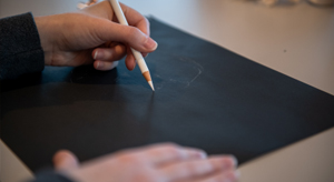 Dispassionate Observation event, a hand holding a white pencil drawing on black paper