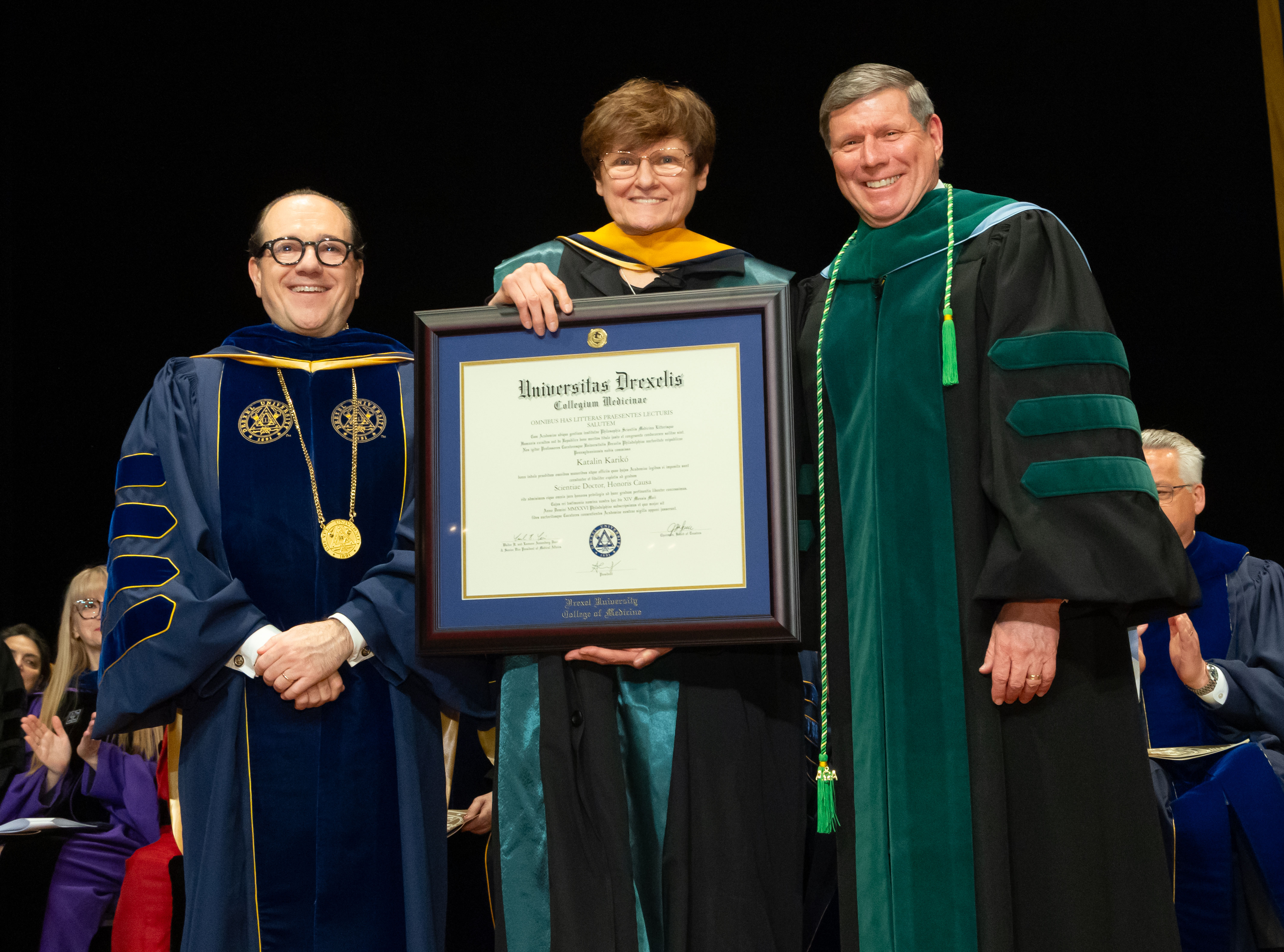 Three people, with center person holding up honorary diploma