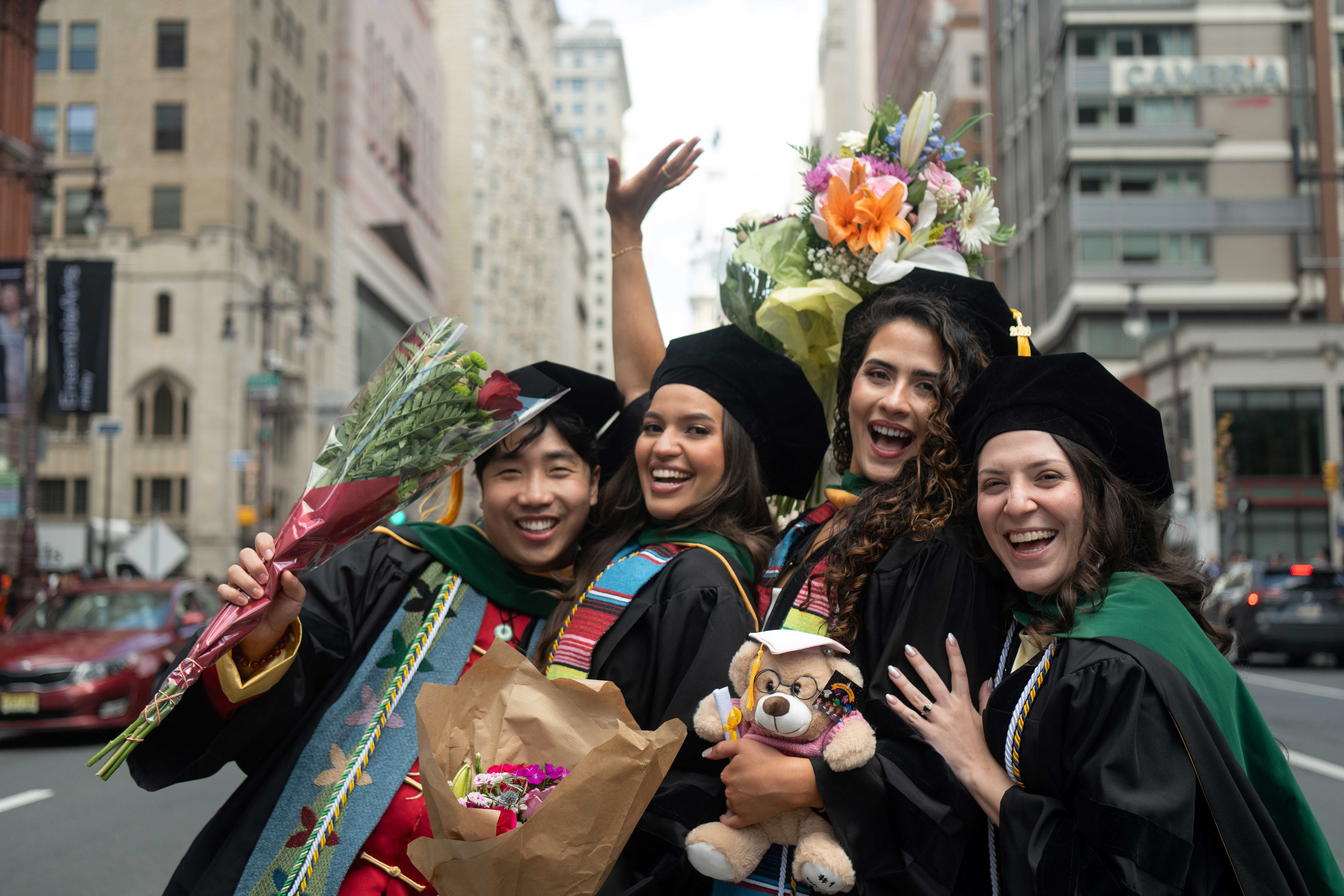four graduates, one raising arm, several holding flowers, a graduate teddy bear on the street