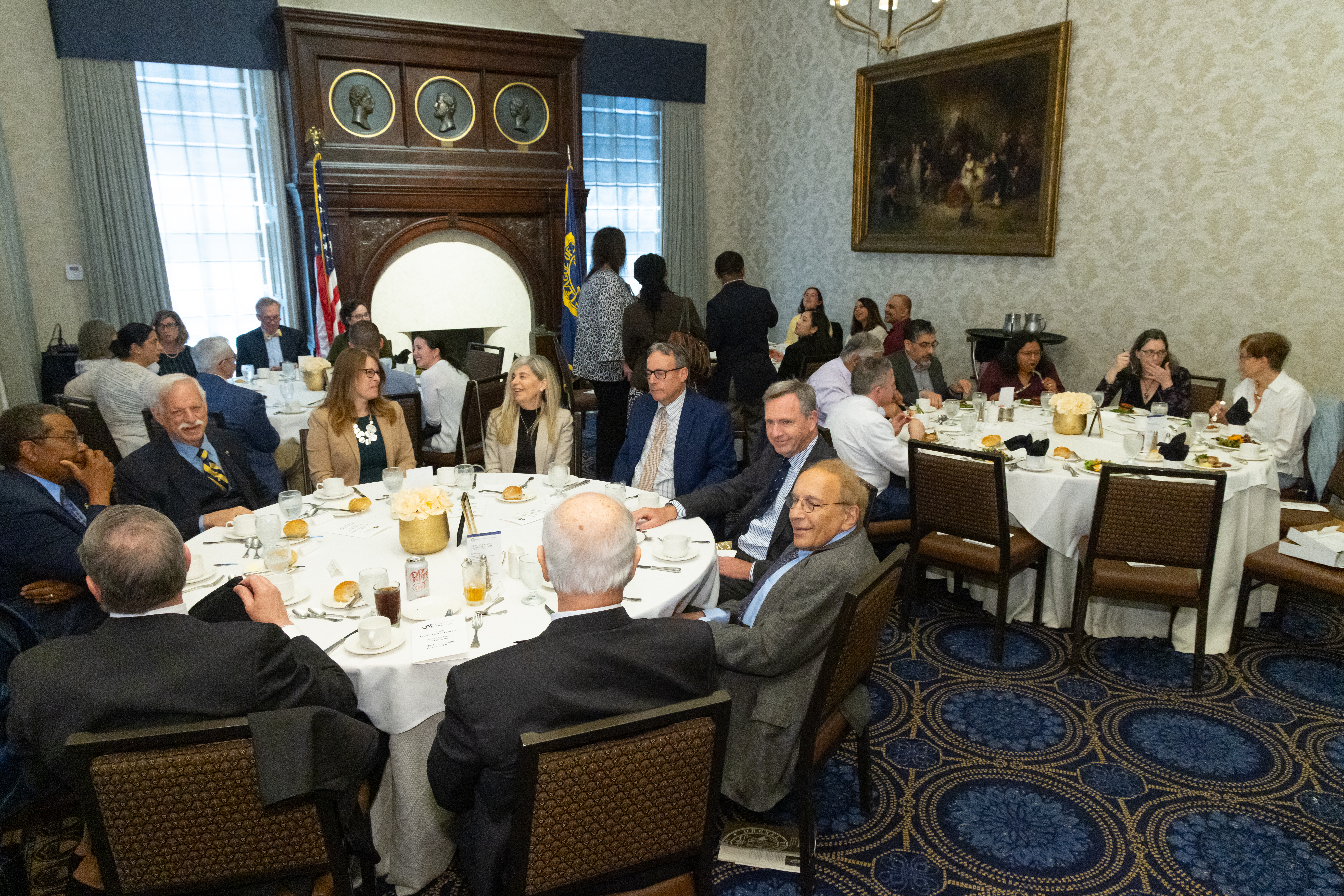 dozens of people sit at round tables while people in background milling about