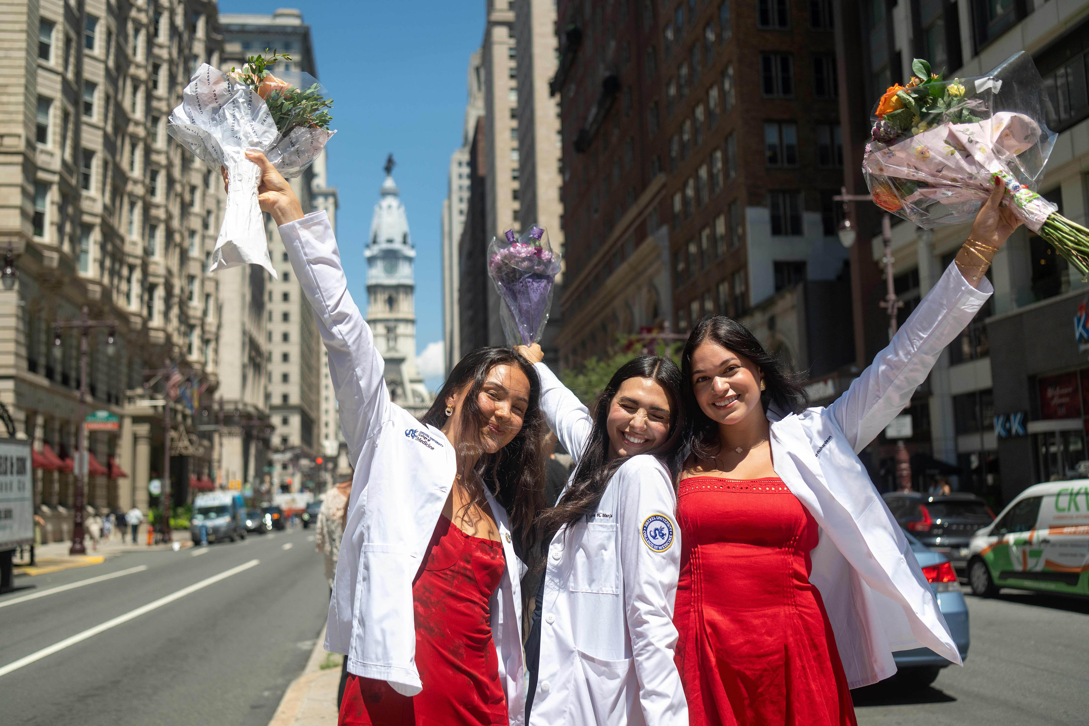 3 students wearing their white coats and raising bouquets of flowers smiling with Philadelphia cityscape behind them.