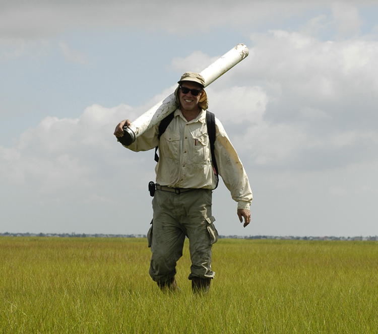 Velinsky standing in a salt marsh with a core barrel over his shoulder