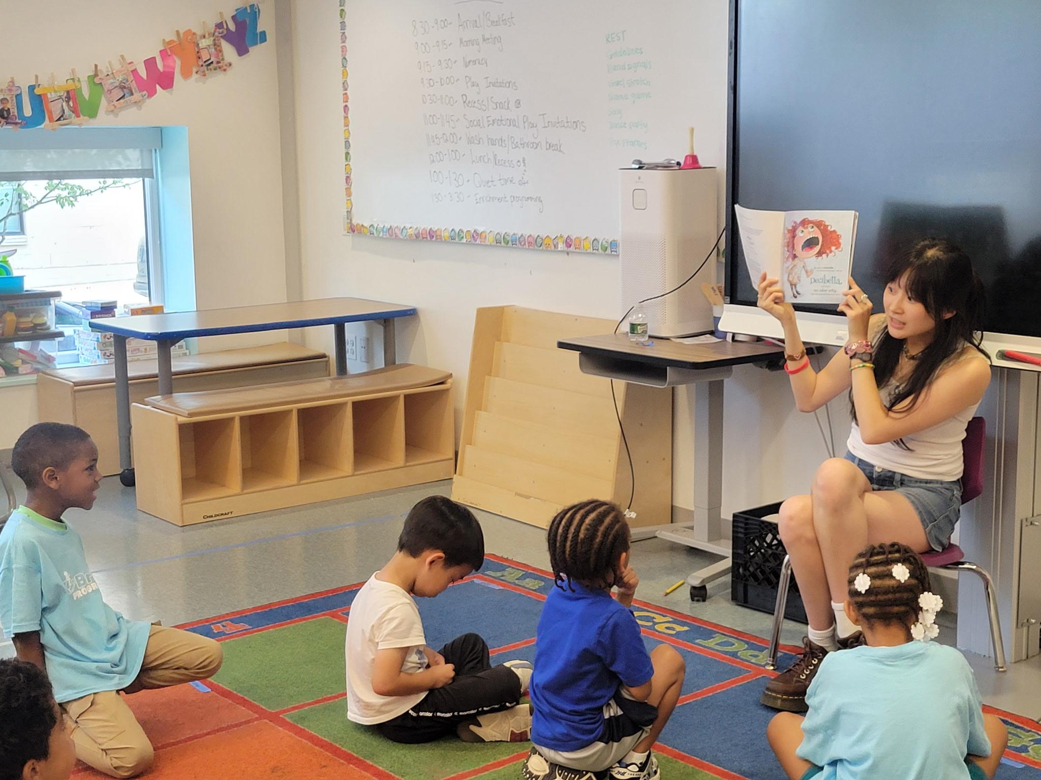 Naomi Yoon reads a picture book aloud to a small group of young children sitting on a colorful classroom rug at Powel Elementary School.