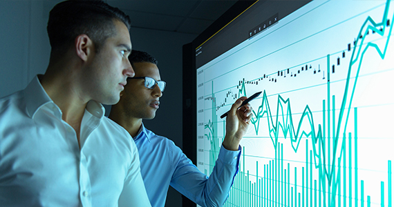 Two men in front of a screen depicting multiple types of graphs and data 